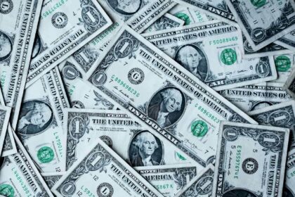 Pile of scattered one-dollar bills showing portraits of George Washington and green seals and serial numbers in a money-filled scene.