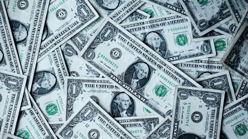 Pile of scattered one-dollar bills showing portraits of George Washington and green seals and serial numbers in a money-filled scene.