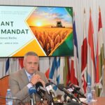 Politician at a press conference seated at a table with multiple microphones, flags in the background and a large screen displaying 'BILANȚ DE MANDAT' behind him.