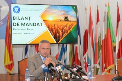 Politician at a press conference seated at a table with multiple microphones, flags in the background and a large screen displaying 'BILANȚ DE MANDAT' behind him.