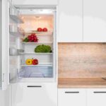 Open refrigerator showing shelves with tomatoes, peppers, greens, and scattered produce in a modern kitchen.