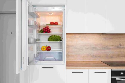 Open refrigerator showing shelves with tomatoes, peppers, greens, and scattered produce in a modern kitchen.