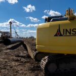 Yellow excavator on a muddy construction site, with a large black pipe nearby and a bright blue sky above.