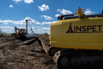 Yellow excavator on a muddy construction site, with a large black pipe nearby and a bright blue sky above.