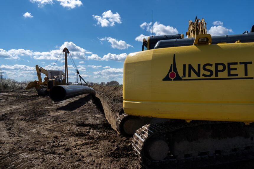 Yellow excavator on a muddy construction site, with a large black pipe nearby and a bright blue sky above.
