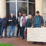 Group of people standing in a line outside a building, holding hunger-strike signs.