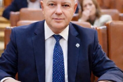 Man in a navy suit and blue patterned tie sits at a wooden desk in a formal meeting room, facing the camera.