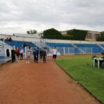 Small group of people stand on a dirt running track beside blue bleachers at an outdoor stadium on an overcast day.