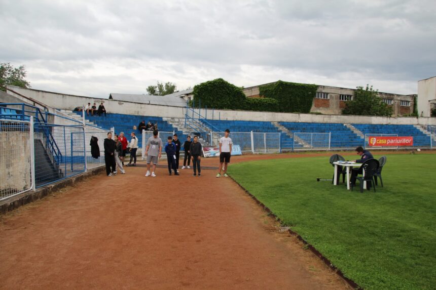 Small group of people stand on a dirt running track beside blue bleachers at an outdoor stadium on an overcast day.