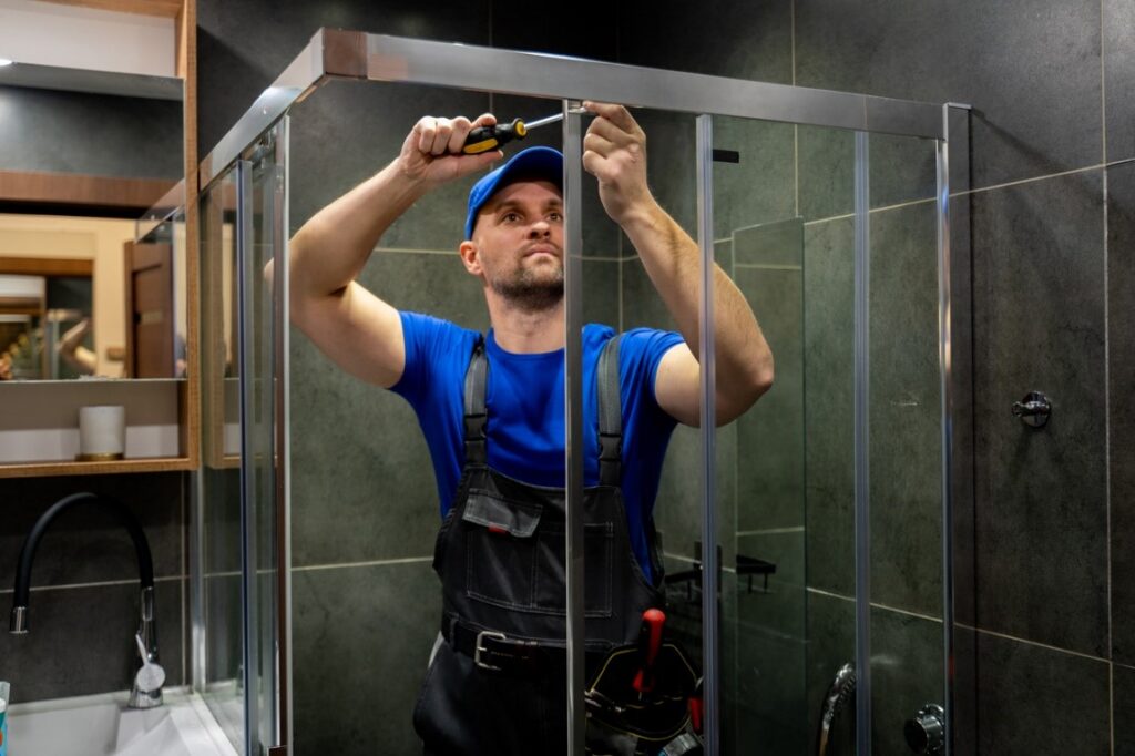 mester-renovare-baie - Jurnalul Olteniei – Știri din Craiova și Oltenia Male handyman in a blue shirt and cap screws a metal frame of a glass shower enclosure in a bathroom using a screwdriver