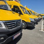 Row of bright yellow vans parked in a row in a lot, with a blue sky overhead and people standing nearby.