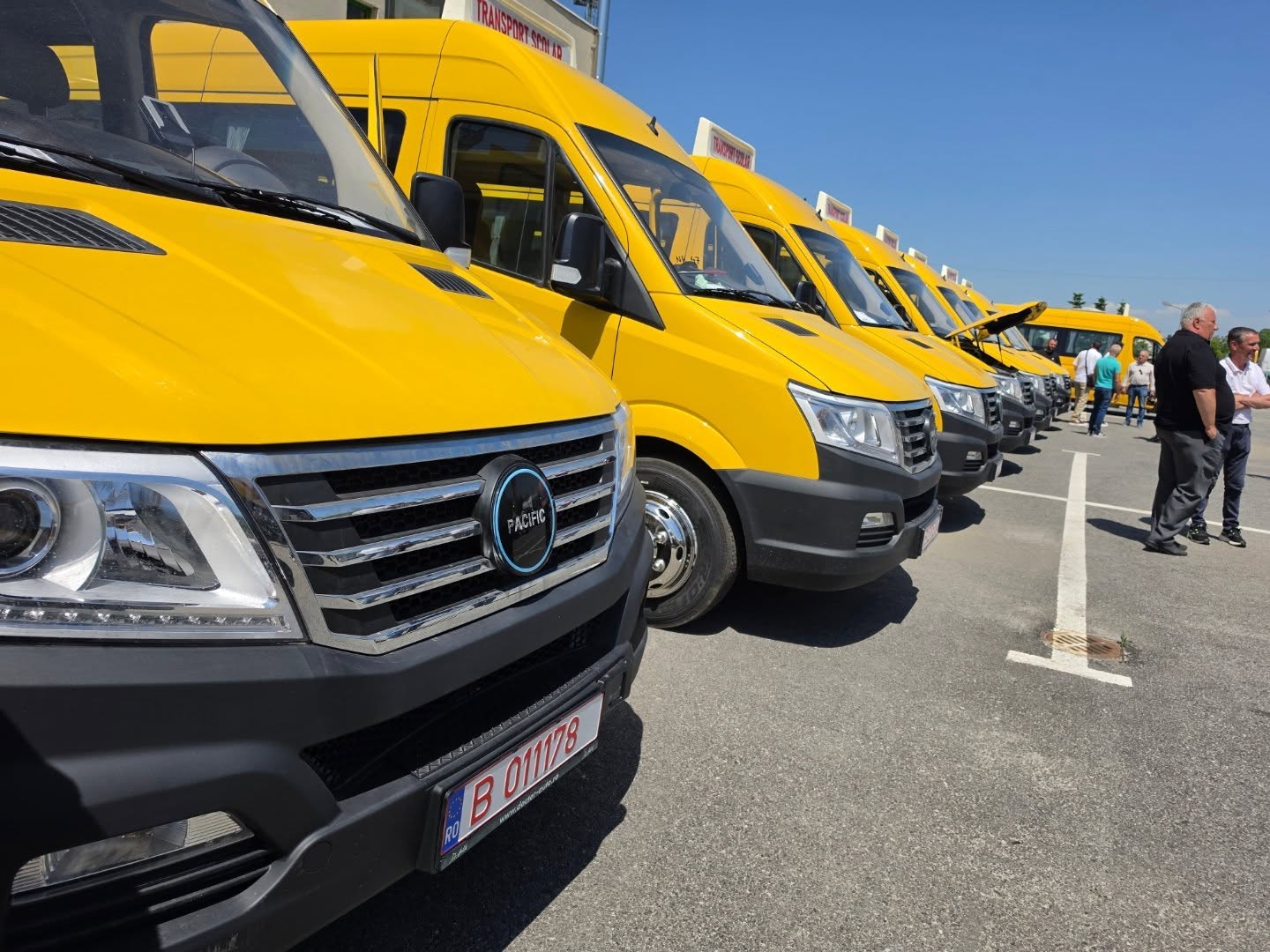 Row of bright yellow vans parked in a row in a lot, with a blue sky overhead and people standing nearby.