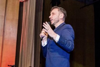 Male speaker in a blue suit delivering a talk on a stage, holding a microphone and gesturing with his hand.