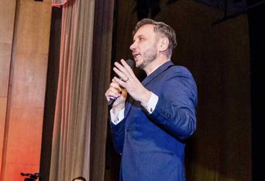 Male speaker in a blue suit delivering a talk on a stage, holding a microphone and gesturing with his hand.