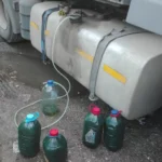 Under a parked truck, two large metal fuel tanks with yellow strips, hoses and caps, and several green jugs with red caps on a muddy ground.
