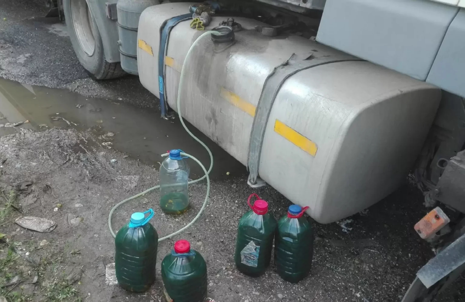 Under a parked truck, two large metal fuel tanks with yellow strips, hoses and caps, and several green jugs with red caps on a muddy ground.