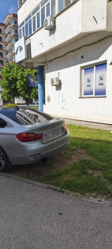 Silver BMW sedan parked on a grassy verge beside a white building with PNL window signs, a blue support column, and wall-mounted air conditioners.