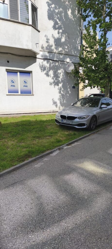 Silver BMW parked on a grassy verge beside a white apartment building, with a satellite dish on the wall and tree shadows nearby.