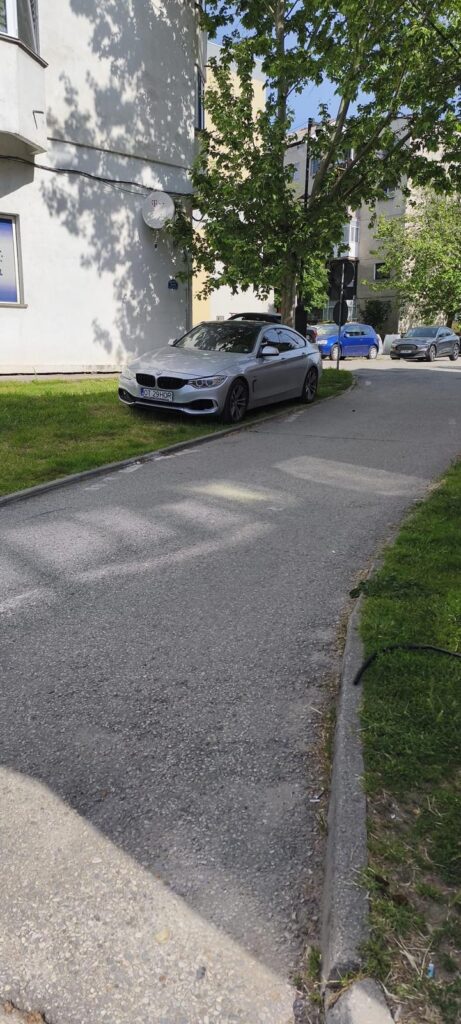 Silver BMW parked on a grassy verge beside a sidewalk in a residential street with trees and apartment buildings in the background.