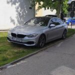 Silver BMW sedan parked on a grassy curb beside a sidewalk, facing slightly left.