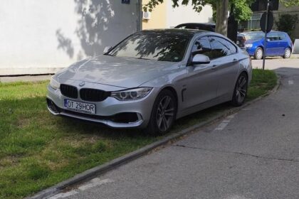 Silver BMW sedan parked on a grassy curb beside a sidewalk, facing slightly left.