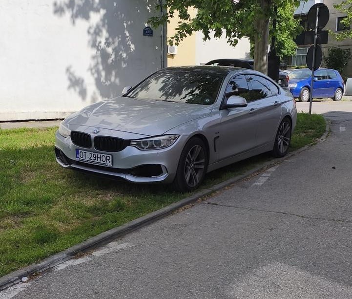 Silver BMW sedan parked on a grassy curb beside a sidewalk, facing slightly left.