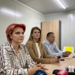 Three professionals sit at a conference table during a press briefing, microphones in front of them and a woman with red hair on the left.