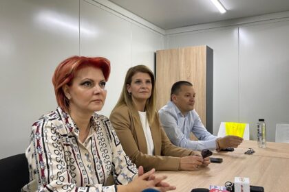 Three professionals sit at a conference table during a press briefing, microphones in front of them and a woman with red hair on the left.