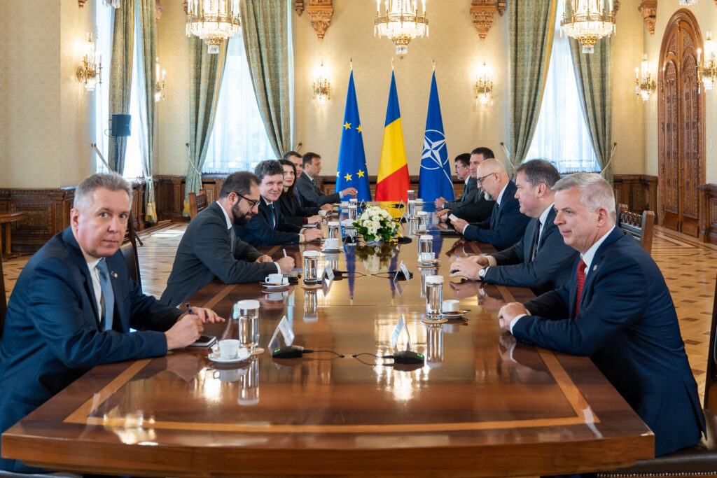 A formal meeting with officials seated along a long wooden table, flags of the EU, Romania, and NATO in the center, ornate chandeliers and curtains in the background.