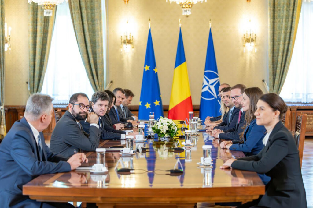 Formal meeting with officials seated on both sides of a long wooden table, flags in the background (EU, Romania, NATO).