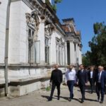 Group of men in business attire walking along the side of an old ornate white building with carved windows under a clear blue sky.