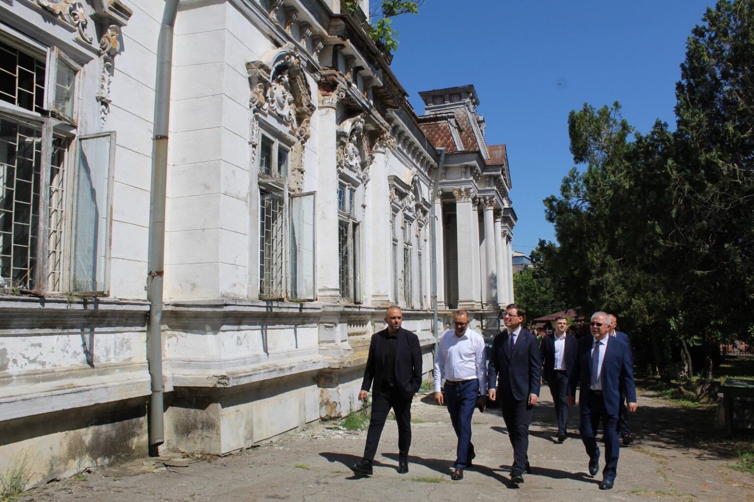 Group of men in business attire walking along the side of an old ornate white building with carved windows under a clear blue sky.