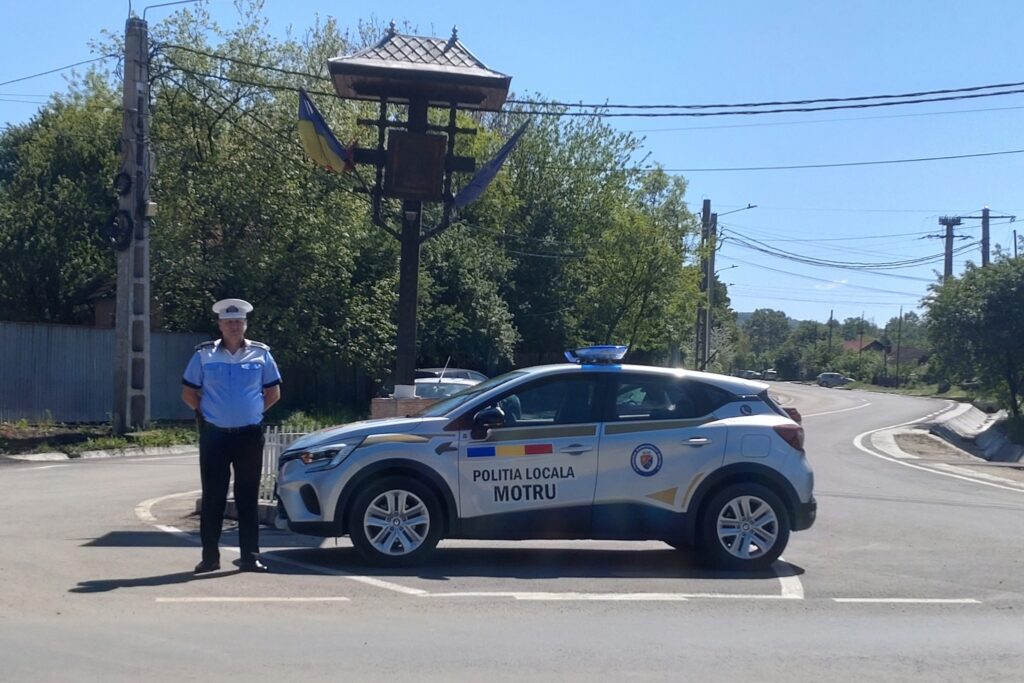 Local police car labeled Politia Locala Motru on a curved road with a uniformed officer standing nearby, sunny day.