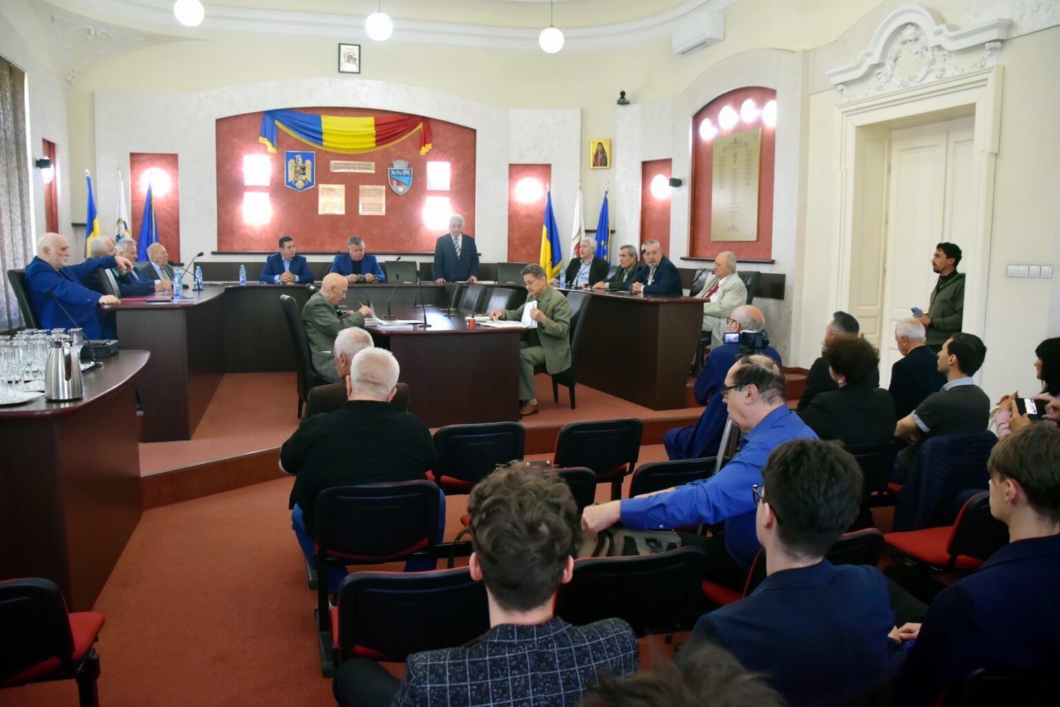 A formal meeting in a large room with a U-shaped table, several men in suits seated around the table, and a speaker standing at the center front while an audience watches from rows of chairs.