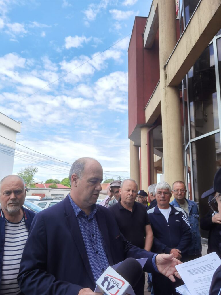A group of men in suits speaking to reporters outside a building, microphones and papers visible in the foreground against a blue sky with clouds
