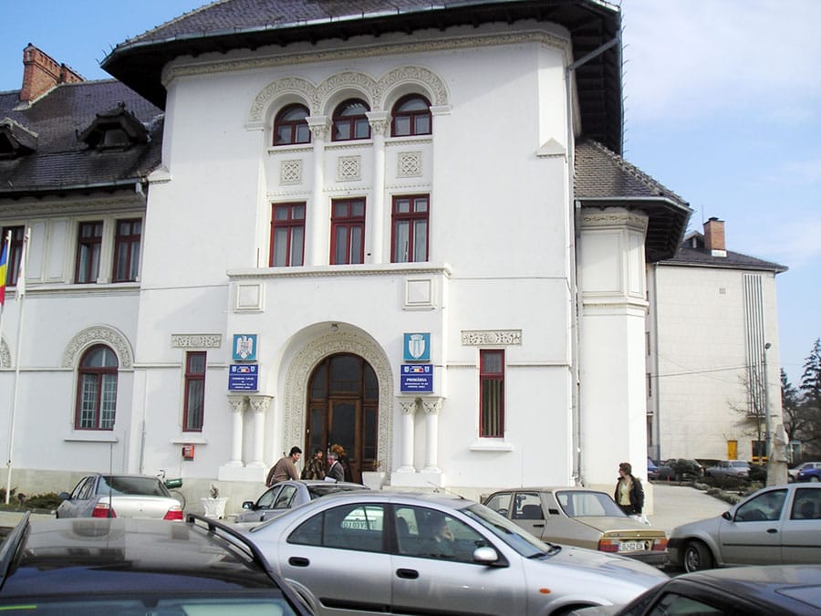 Front of a white historic building with arched windows and decorative facade, blue signs by the entrance, and cars parked outside.