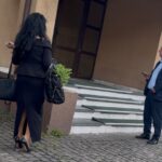 Two professionally dressed people at a building entrance: a woman in a black outfit with high heels carrying bags, facing away; a man in a suit stands on the steps looking at his phone.