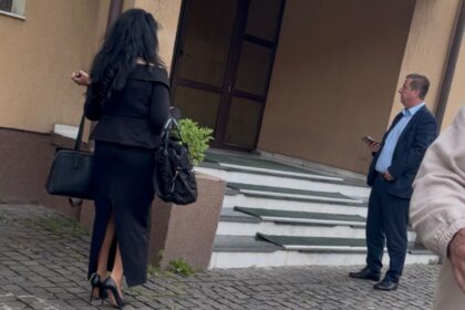 Two professionally dressed people at a building entrance: a woman in a black outfit with high heels carrying bags, facing away; a man in a suit stands on the steps looking at his phone.