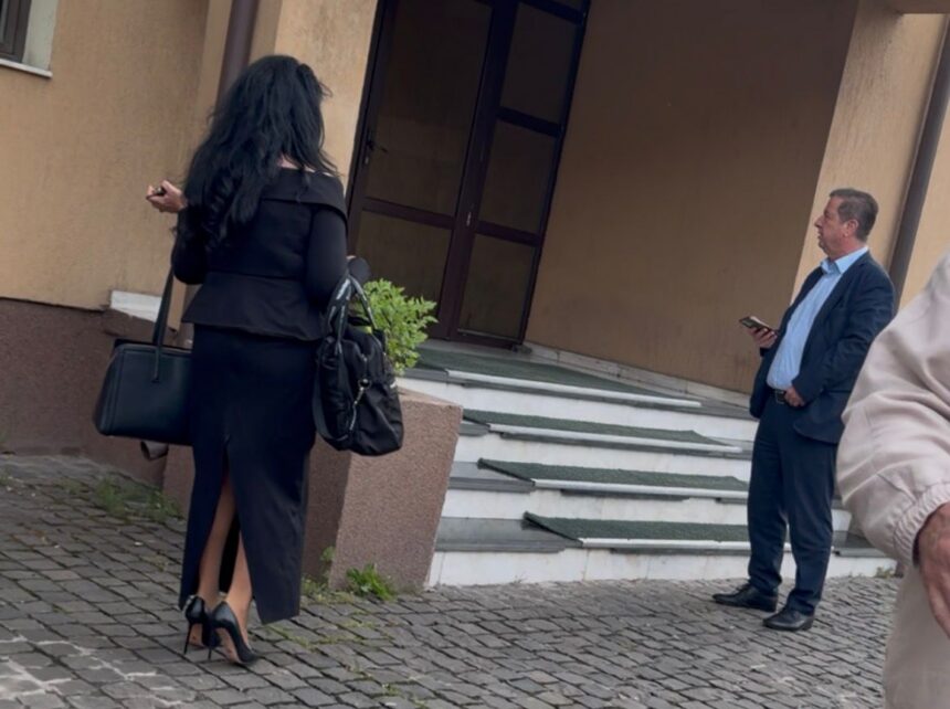 Two professionally dressed people at a building entrance: a woman in a black outfit with high heels carrying bags, facing away; a man in a suit stands on the steps looking at his phone.
