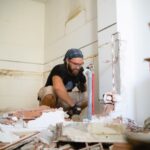 Construction worker with a bandana chiseling through bricks in a partially demolished room filled with rubbleing debris on the floor