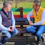 Two men sit on a colorful park bench outdoors, talking in a casual interview.