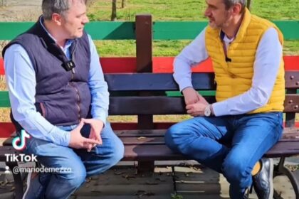 Two men sit on a colorful park bench outdoors, talking in a casual interview.
