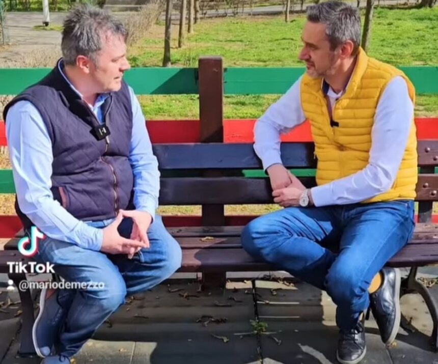 Two men sit on a colorful park bench outdoors, talking in a casual interview.