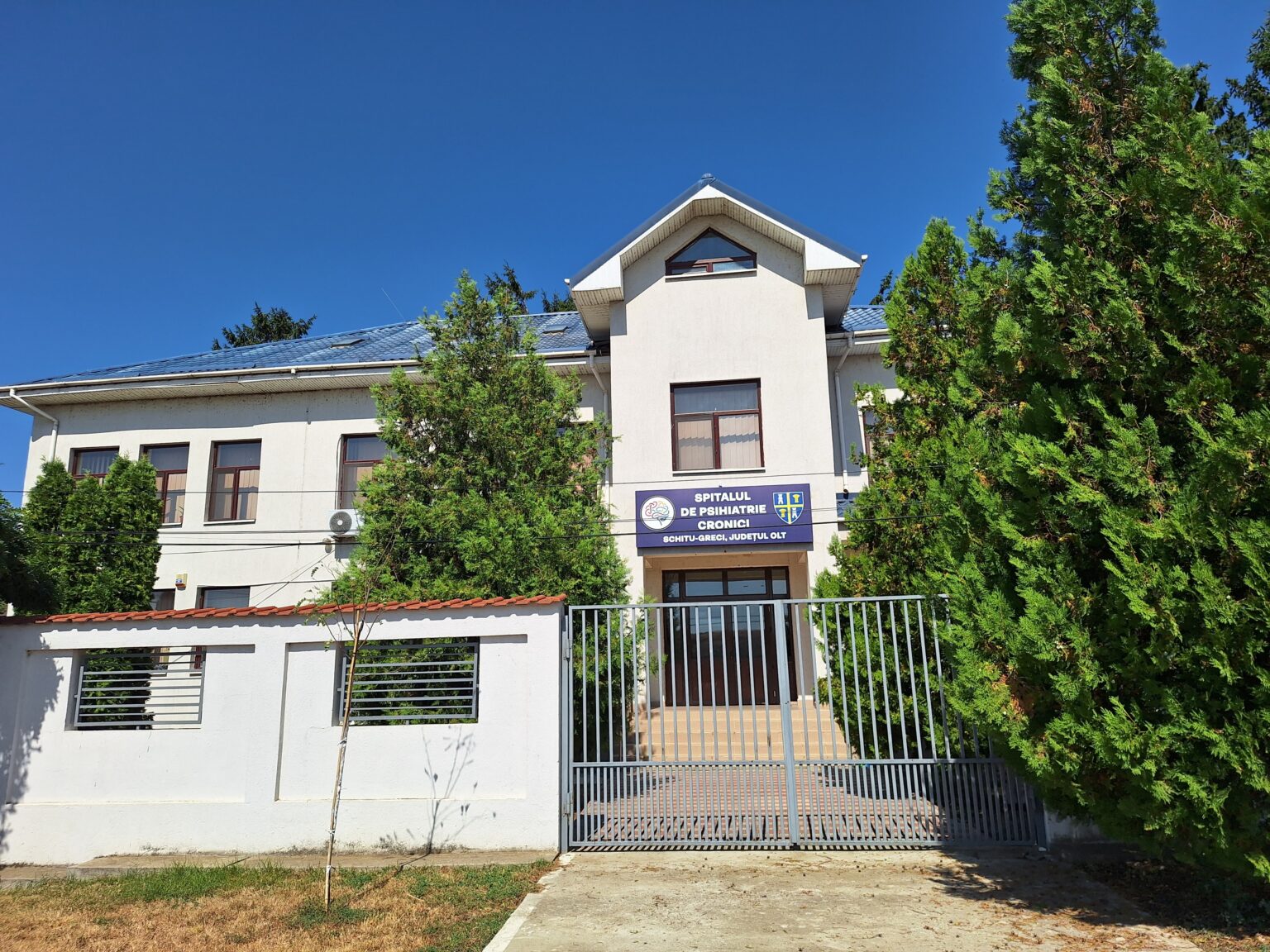 Hospital building with a blue sign in Romanian, flanked by trees and a metal gate under a clear blue sky