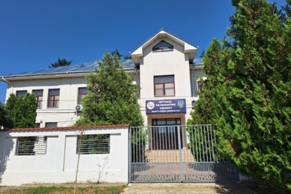 Hospital building with a blue sign in Romanian, flanked by trees and a metal gate under a clear blue sky