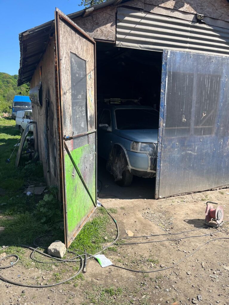 Weathered shed with an open door revealing a silver-blue car inside; cluttered yard with hoses and equipment outside.