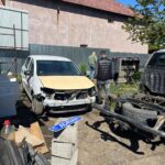 Damaged white car with a missing front bumper in a junkyard, two men inspecting it near scattered auto parts and a sign reading 'ADRIAN' on a concrete block.