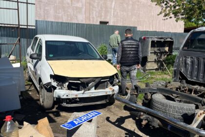 Damaged white car with a missing front bumper in a junkyard, two men inspecting it near scattered auto parts and a sign reading 'ADRIAN' on a concrete block.