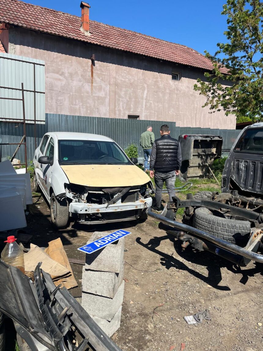 Damaged white car with a missing front bumper in a junkyard, two men inspecting it near scattered auto parts and a sign reading 'ADRIAN' on a concrete block.