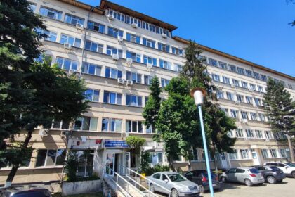 Facade of a large beige, multi-story hospital/official building with many windows, entrance with a ramp, parked cars, and surrounding trees under a clear blue sky.